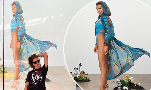 Irina Shayk poses in front of a poster of herself flaunting her jaw-dropping physique in a racy blue swimsuit and a floral hooded cape for Ivy Park shoot