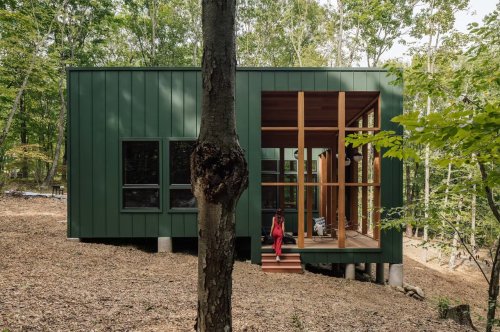 This dark green cabin floats above a sloping terrain in a forest in Connecticut